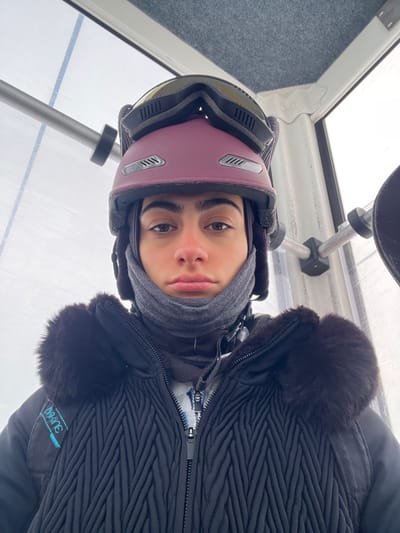 A single individual is depicted in a close-up, chest-up perspective within a ski gondola cabin located in Valtournenche, Italy. The subject wears a dark maroon ski helmet, topped with dark-framed ski goggles featuring yellow-tinted lenses. A grey neck gaiter covers the lower face, exposing the nose and a neutral-expression mouth. The subject's nose exhibits mild redness. Brown eyes gaze directly forward beneath dark, prominent eyebrows. Outerwear consists of a black winter jacket, characterized by a chevron-patterned quilted front and dark fur trim around the collar. A blue and white patterned garment is partially visible beneath the unzipped jacket. A black strap, possibly from a backpack, bearing a light blue 'DAKINE' logo, is visible on the left side of the frame. The background confirms an indoor setting within the gondola cabin, featuring a grey, textured ceiling and windows looking out onto a bright, snow-covered winter environment. Metal support bars are present within the cabin's interior structure.