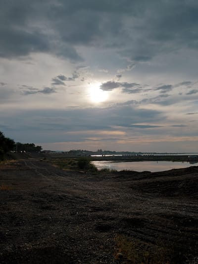 The image depicts a landscape scene near the city of Ақши, Kazakhstan, characterized by a cloudy sky with the sun partially visible and peeking through. The foreground is dominated by rough, dark terrain, appearing to be dirt and gravel, possibly a construction site or an unpaved road. To the left, a dirt track winds upwards, flanked by sparse vegetation and trees. Beyond this, a line of cars is parked along a raised embankment. In the middle ground, a body of water, likely a lake or river, stretches across the scene, reflecting the sky. Along the far bank of the water, a cluster of buildings and structures can be seen, suggesting a settlement or industrial area. A lone car is visible on the far right. The overall impression is one of a natural and developing landscape under a moody sky.