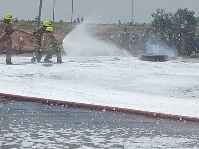 Three firefighters wearing yellow helmets are standing in a field covered in white foam. The firefighters are holding a hose and spraying the foam onto a burning object in the distance. The firefighters are standing on the edge of the foam, and the foam is spread out over a large area. The location of the scene is Katabu, Nigeria, at coordinates 10.71 latitude and 7.514 longitude. The sky is overcast and there are raindrops falling. There are several people in the background, but they are not visible in detail. The scene is a training exercise for the fire department.