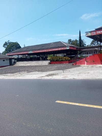 This image depicts an outdoor scene featuring a food establishment, viewed from across a paved road under a clear blue sky.
The primary subject is a multi-tiered restaurant or food stall complex, identifiable by red banners prominently displaying the Indonesian text "TAHU SUMEDANG RENYAH" (Crispy Sumedang Tofu) along the perimeter of its dark-colored roof. This linguistic detail indicates the location is in Indonesia, not Beteng_Pangkhar_Somthrang, Bhutan, as specified.
In the foreground, a dark gray asphalt road runs horizontally across the bottom third of the frame. A short, dashed yellow lane marking is visible on the right side of the road. The road appears dry and devoid of traffic or pedestrians.
Behind the road, a lighter gray, uneven ground surface, possibly a parking area, extends towards the restaurant. Two slender, metallic posts are vertically embedded in this ground. To the right, a more structured paved area with some surface cracks leads towards a set of red-painted concrete stairs that ascend to a higher section of the complex.
The main restaurant structure features an open-air design with visible tables and chairs, some of which are white plastic. Green foliage, likely ornamental plants, is arranged in a long red planter box in front of the elevated dining area. To the far left, a smaller, single-story white building with a dark roof is partially visible, bearing a sign indicating "WC." Large green trees form a backdrop to the complex, with one particularly tall, slender tree rising centrally behind the main building.
The scene is brightly illuminated by natural daylight, suggesting mid-morning or early afternoon. No individuals are visible within the frame. The overall impression is that of a quiet, operational roadside eatery.