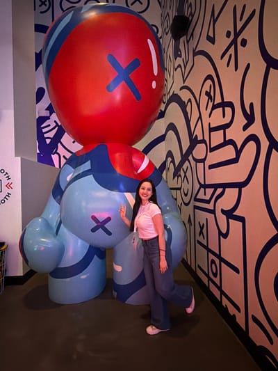 An indoor scene in Las Vegas, United States of America, depicts a young adult female posing next to a large, stylized inflatable figure. The female, with long dark hair, wears a light pink crop top, dark flared jeans, and white sneakers, and has an ID badge on a lanyard around her neck. She stands to the right of the figure, smiling towards the camera with her right hand raised in a peace sign. The inflatable figure is predominantly light blue and red, featuring a large red spherical head with a dark blue 'X' mark, and a bulbous light blue body with another dark blue 'X' mark on its rear. Dark blue stripes and smaller light blue accents adorn its form. The background consists of a wall covered in a dense, dark blue doodle-style mural on a light beige surface, featuring abstract shapes, 'X' marks, circles, and arrows. To the far left, a light gray wall section displays vertical text, possibly "BOOTH," and a metal wire basket is partially visible. The floor is dark and uniform.