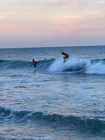 The image depicts a serene coastal scene at Arugam Bay, Sri Lanka, during what appears to be a sunset or sunrise, evidenced by the soft pastel hues of pink and blue in the sky. The ocean is calm, with gentle waves rolling in. In the foreground, the water is a deep blue, with white foam cresting on the waves.
In the mid-ground, a surfer is actively riding a wave. The surfer, appearing to be male, is positioned on a light-colored surfboard, demonstrating skill as they navigate the curl of the wave. The surfer is shirtless, wearing light-colored shorts. To the left of this surfer, further back, another individual is visible on a surfboard, wearing a bright orange vest, indicating they may be a beginner or engaging in a water activity that requires higher visibility. Another individual is further in the background, also on a surfboard. The overall environment suggests a popular spot for surfing and other water sports.