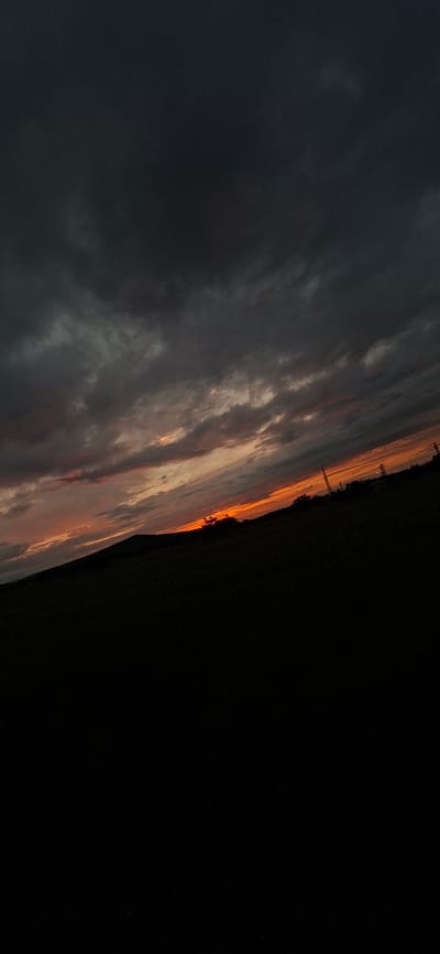 The image captures a dramatic twilight sky over a dark, silhouetted landscape, likely a rural setting near Lyaskovets, Bulgaria. The sky is the dominant feature, displaying a striking contrast between dark, heavy clouds in the upper left and vibrant warm colors of orange, red, and gold along the horizon on the right. These luminous hues suggest either a sunset or sunrise, with the sun's low angle illuminating the edges of the cloud formations.

Below the colorful sky, the foreground appears as a deep, almost black silhouette, representing fields, distant trees, or hills. Along the jagged horizon line, faint outlines of what appear to be electricity pylons or communication towers are visible against the brighter parts of the sky, providing a subtle hint of human infrastructure. There are no people, explicit activities, or visible text in the image. The weather appears partly cloudy to overcast, contributing to the dramatic atmosphere. The scene evokes a sense of tranquil beauty and the profound spectacle of nature during the fleeting moments of dawn or dusk.