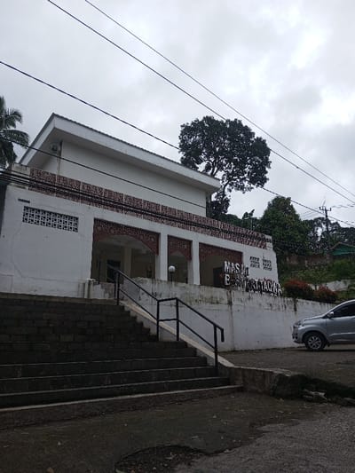 The image displays the "MASJID BAITATTAQWA" building in Kodarapalli, India, under an overcast sky. The two-story structure features white walls, a flat roof, and decorative reddish-brown patterns above the ground-level entrance arches and along a horizontal band. A loudspeaker is mounted on the upper left corner of the facade. Access to the building is provided by a wide, dark grey stone staircase equipped with black metal handrails, leading up to the main entrance. A white retaining wall supports the building, with "MASJID BAITATTAQWA" inscribed in dark letters on its surface to the right of the stairs. A silver compact SUV is parked on a wet, paved surface at the base of the retaining wall. Lush green vegetation, including large trees with dense canopies, is visible behind and to the right of the building, with red flowering bushes along the retaining wall. Multiple utility wires cross the upper portion of the frame. No individuals are present in the scene.