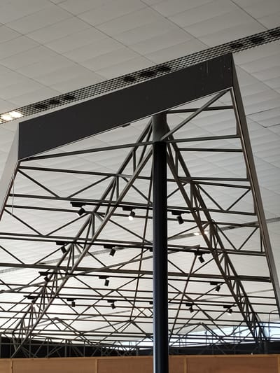 The image shows a view looking up at a large, dark, rectangular sign structure supported by a complex network of metal beams and struts. The sign itself is dark and appears to have some scuffs or marks on its surface. The metal framework is triangular and extends upwards, with multiple cross-braces and diagonal supports. Several small, dark spotlights are attached to the beams. Above the sign and its supporting structure, a white ceiling with a grid pattern is visible. The lower part of the image shows a wooden paneling or counter. This scene is likely within a large public building such as an airport terminal or exhibition hall. The reference to "el Prat de Llobregat, Spain" strongly suggests this is a view within Barcelona-El Prat Airport, a major international airport serving the city of Barcelona and the surrounding region.