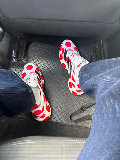 An overhead view captures the lower body of an individual seated within a vehicle. The subject wears dark blue denim jeans and white athletic shoes patterned with large red polka dots, featuring black Adidas stripes on the sides. White laces and white socks are visible. Their feet rest on a black rubber floor mat characterized by a raised diamond plate texture. A black strap with a plastic buckle lies on the floor mat to the right of the right foot. A dark car seat with light grey stitching is partially visible in the lower foreground. The exact location within Hazel Grove, United Kingdom, is not discernible from the image content.