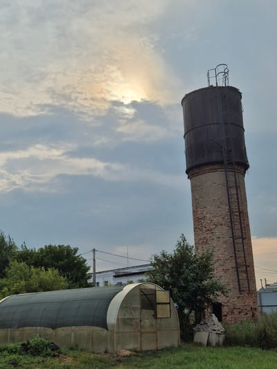 The image shows an exterior scene in Новоишимское, Kazakhstan, featuring a tall, weathered brick water tower with a dark, cylindrical top and an attached metal ladder. The tower is situated in a grassy area with some foliage. To the left of the tower, a large, semi-circular greenhouse with a dark mesh covering and translucent panels is visible. In the background, there are trees, a white building, and utility poles with wires. The sky is overcast with patches of sunlight breaking through the clouds. There are no people present in the image.