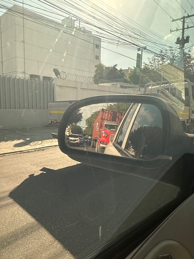A car's side mirror reflects a red truck followed by a white pickup truck. Outside the car, a modern building with many windows is visible. There's a fence topped with barbed wire and telephone poles with wires are in view. The road is asphalt with a shadow cast on it. The image is taken from inside a car, capturing the interior door and window.