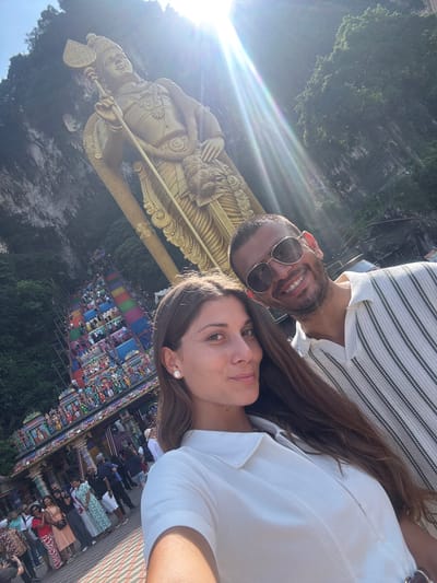 A couple poses for a selfie with a large golden statue of Lord Murugan at the Batu Caves in Selayang Municipal Council, Malaysia. The man, wearing sunglasses and a striped shirt, is smiling, as is the woman, wearing a white shirt. The statue is in the foreground. The background features the colorful steps and the surrounding greenery of the Batu Caves complex. Sunlight and lens flare are visible in the upper left corner.