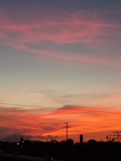 The image depicts a dramatic sunset or sunrise over a silhouetted landscape, set in El Dátil, Venezuela. The sky dominates the scene, displaying a striking gradient of colors. At the top, the sky is a deep purple-blue, transitioning downwards through soft pink and lavender hues, culminating in vibrant oranges and fiery reds near the horizon. Streaky, elongated clouds are visible across the expanse, reflecting the intense colors of the low-lying sun.
The lower part of the image features a dark silhouette of the terrain, indicative of strong backlighting. Several utility poles, one prominently positioned in the mid-ground, stand against the colorful sky, with overhead power lines stretching horizontally across the frame. Below the horizon line, faint outlines of what appear to be vehicles with their lights on are visible on a road, suggesting activity during this twilight period. No people are discernibly visible, and there is no visible text in the image. The overall mood conveyed is serene and dramatic, capturing a fleeting moment of natural beauty.
