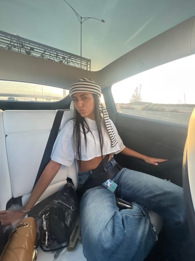 The image shows a young woman seated in the rear passenger side of a vehicle in Kafr Kasim, Israel, during daylight hours, likely late afternoon. The woman, who has long dark hair, is wearing a white and black striped headscarf, a white short-sleeved crop top, and loose blue denim jeans. A black seatbelt is fastened across her chest and lap. She is looking directly at the camera with her lips pursed. Her left arm rests near a black quilted shoulder bag with a silver chain strap positioned on the white seat beside her. Her right hand is placed on the interior door panel. A light-colored card or tag is visible tucked into her jeans under the seatbelt on her right side. The vehicle's interior features white seating and a panoramic glass roof. Outside the vehicle, visible through the windows, is a multi-lane roadway. On the left, a large bridge structure with metal railings is visible, while on the right, sparse vegetation, including trees and utility poles, is present under a bright sky with a low sun.