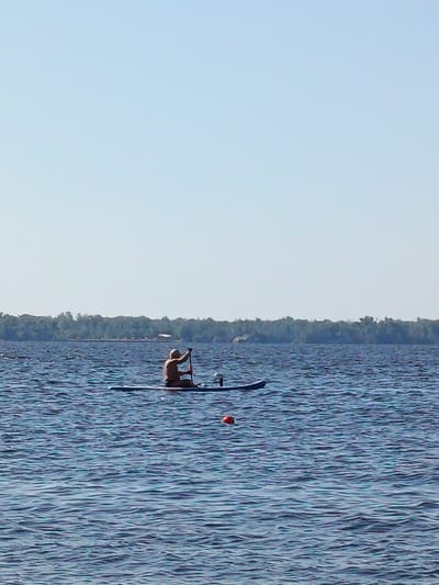 A person is paddleboarding on a body of water with a forest line in the background. The person is sitting on the board and holding a paddle. There is a red buoy in the water to the right of the paddleboarder. The sky is clear and blue. The location is Eremeevka, Russia.