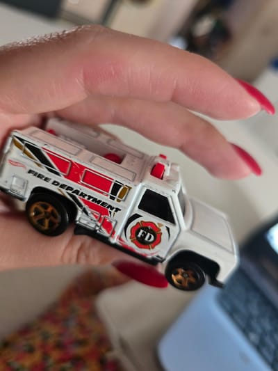This is a close-up, eye-level shot of a person's hand holding a miniature white fire truck. The hand has long, well-manicured red nails. The fire truck is detailed with red and black accents, and the words "FIRE DEPARTMENT" are clearly visible along its side in black lettering. The "FD" logo is also present on the side of the truck. The fire truck appears to be a toy, likely a Hot Wheels model, as the logo is visible at the bottom left of the vehicle.
The background is out of focus, but hints of a laptop and a colorful patterned fabric can be seen, suggesting an indoor setting. The lighting is warm, possibly indicating daytime or interior lighting. No specific weather or time of day is apparent beyond this. The overall mood is casual and focused on the toy vehicle. The location context of Petropavlivska Borshchahivka, Ukraine, is noted but not visually indicated in the image itself.