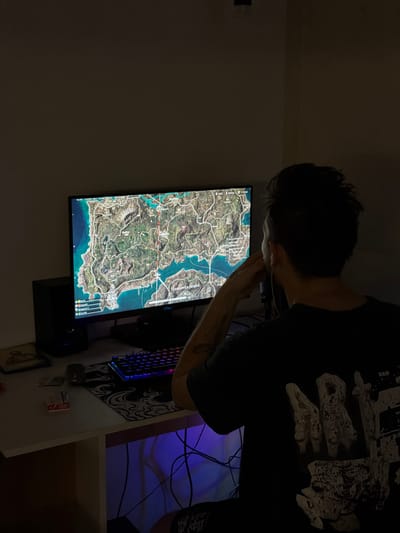A person stands, hands clasped near their face, intently observing a computer monitor displaying a detailed geographical map. A gaming keyboard, adorned with illuminated keys, sits on a patterned mousepad. Behind the desk, a blue light casts a faint glow. Wires are visible tangled behind the desk. The overall lighting is dim, casting shadows around the room.
