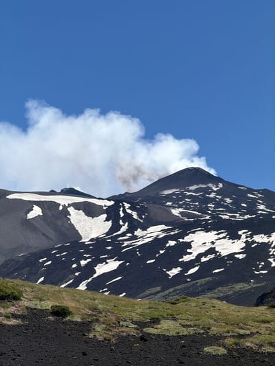 A panoramic, low-angle, outdoor, vertical shot captures Mount Etna, a stratovolcano located near Linguaglossa, Italy, on a bright, clear day. The sky is a vibrant, cloudless blue, with a few wisps of white cloud near the peak of the mountain. The volcano's dark, rocky slopes are partially covered in patches of white snow, creating a stark contrast. A plume of grey smoke emanates from the volcano's summit, indicating volcanic activity. The foreground of the image features a dark, rocky terrain covered with sparse green grass and small bushes. The lighting suggests it is daytime, with shadows cast on the mountainside. No people are visible in the image.