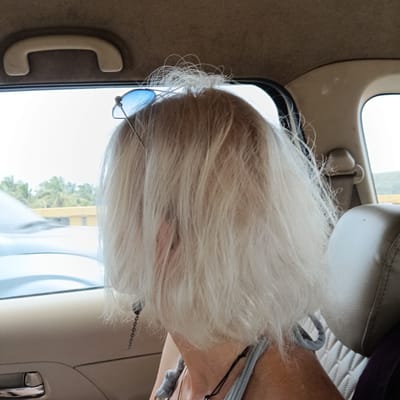 The photograph, taken in the interior of a vehicle, possibly a car, in Chopdem, India, shows the back of a person's head and shoulders. The individual has short, layered, platinum blonde hair with sunglasses perched on top. The hair obscures much of their face. The person is wearing a light gray tank top and a necklace with a black cord. A silver chain, potentially an earring or ornament, hangs near their left shoulder. The vehicle interior is beige and gray, with a window partially visible, showing a blurred view of what appears to be another car and some tropical trees, suggesting a warm climate. A beige grab handle is visible on the ceiling of the vehicle.