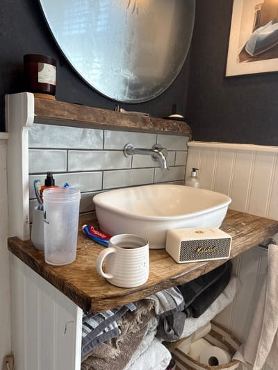 The image depicts a bathroom interior. A round mirror is mounted on a dark wall to the left, and a vessel sink sits above a countertop with light gray tile. A chrome faucet is positioned next to the sink. Adjacent to the sink, on the countertop, are various items including a white Marshall speaker, a striped mug, a tube of Colgate toothpaste, and a translucent shaker bottle containing toothbrushes. A lit candle rests on the corner of the countertop closest to the mirror. A wooden structure holds the countertop and also acts as a shelf, where towels and toilet paper are stored in a wicker basket. The style suggests a rustic or farmhouse aesthetic. While the image does not definitively confirm the location, it is a bathroom interior that could plausibly be in a residential or commercial setting in Hitchin, United Kingdom, based on stylistic cues.