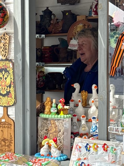 The image shows an elderly woman peering into a display case filled with various Russian souvenirs and crafts, seemingly in Arzamas, Russia. The display contains numerous decorative items, including painted wooden cutting boards, a traditional Russian doll, and ceramic figurines of geese with "Арзамас 1578" printed on them, referencing the city's founding year. A St. George ribbon is visible at the top of the frame. The woman is wearing a blue shirt and appears to be interacting with the items in the case, possibly a vendor showing her wares.