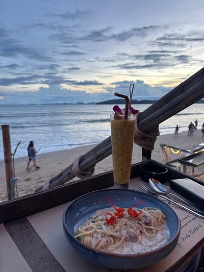 At a beachfront establishment in Ban Klong Haeng, Thailand, a meal is presented for consumption. In the foreground, a blue bowl contains a serving of pasta with a creamy sauce, garnished with orange flower petals. To the right of the bowl are silverware, including a fork and spoon, and a small black object, possibly an ashtray. Behind the food, a tall glass holds a yellow beverage with small dark flecks, adorned with a purple flower and two straws, one brown and one clear. This dining setup is positioned on a wooden tray, which is angled to reveal the ocean, beach, and sky in the background. The sea is calm, with gentle waves lapping onto the sandy shore. Several individuals are visible on the beach; a couple walks near the water's edge, and other figures are scattered further down the shore. In the distance, a faint outline of hills or landmasses can be seen against the horizon. The sky displays a twilight palette of blues and grays, with clouds suggesting the lingering light of sunset or the approach of night. A wooden structure, potentially part of the restaurant's seating or architectural design, frames the scene from the right.