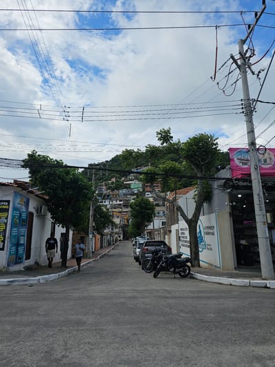 This is a street-level photograph captured in Arraial Do Cabo, Brazil, depicting a sloping urban environment under a partly cloudy sky. The perspective is looking down a street that ascends towards a densely populated hillside.
In the foreground, the street surface is asphalt, with white-painted concrete curbs defining the sidewalks on both sides. On the left sidewalk, two adult males are walking towards the viewer. One wears a dark t-shirt with a light logo and light shorts; the other wears a light t-shirt and dark shorts, holding an item in his right hand. Adjacent to them on the left are white-walled buildings, some with reddish-brown roof tiles. One building features a blue-and-white sign with Portuguese text, including "SUPER PROMO" and "TURISTA" references. An air conditioning unit is visible on a building facade.
On the right side of the street, multiple vehicles are parked along the curb, including a dark gray pickup truck and two motorcycles – one dark-colored with chrome accents and another dark blue/black. Behind the parked vehicles are commercial buildings. One prominent building on the right features a brightly colored pink sign with a ship graphic and Portuguese text, indicating a retail establishment. Another building section has a white wall with a blue and white mural/sign displaying a phone number "22 999441539" and "jetskiarraial". The storefronts show various goods displayed for sale.
Numerous utility poles made of concrete are visible throughout the scene, supporting an extensive and complex network of overhead power lines and communication cables that crisscross the sky. One tall pole on the immediate right features multiple insulators, conduits, and bundles of wires. Several green-leafed trees are interspersed along the street, partially obscuring buildings and power lines.
The background is dominated by a steep, verdant hill covered with numerous residential structures. These buildings, varying in color and construction, appear to be densely clustered, characteristic of hillside communities. The sky above is a mix of blue patches and scattered white clouds.