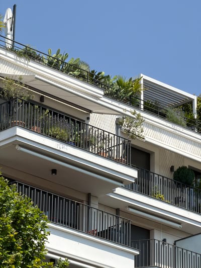 The image depicts a modern apartment building with multiple balconies filled with lush greenery, including cacti, palm trees, and lavender. The architecture is sleek and contemporary, with white facades and black metal railings. The balconies are adorned with potted plants and some appear to have retractable awnings. The building is situated under a clear blue sky, suggesting a sunny day. The location is identified as Neuilly-sur-Seine, France. There are no people visible in the image. The scene is tranquil and urban, showcasing a harmonious blend of modern living and nature.