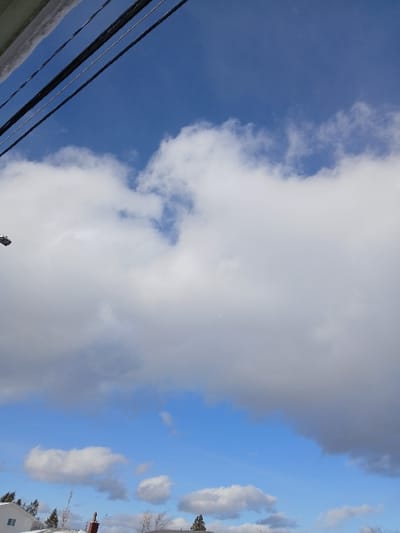 The image shows a sky filled with clouds. On the left, the sky is a deep blue, with fluffy white clouds gradually transitioning to larger, gray clouds that dominate the upper portion of the image. Power lines are visible in the lower left corner. On the right side of the image, the sky is a lighter blue with smaller, scattered clouds. A few trees and a building with a red chimney are visible on the bottom-right of the frame.