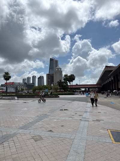 This image shows a city square with a prominent sky filled with dramatic, dark storm clouds and patches of blue sky. In the background, a cluster of modern skyscrapers rises, including a tall, slender building with a distinctive angular top, and a more traditional building with a clock face. A large building with a long, overhanging roof, likely a public transportation terminal, dominates the right side of the frame. The foreground is a wide, paved plaza with a patterned surface of light and dark square tiles, with some areas appearing wet, suggesting recent rain. A single person is riding a small bicycle in the middle ground. Further back, two young men walk together along the plaza, and a crowd of people with umbrellas are visible near the entrance of the large building. Palm trees are scattered throughout the scene, indicating a tropical or subtropical climate. The overall impression is of a busy urban environment on a day with uncertain weather. The city of Xiangzhou, China, is known for its modern architecture and coastal location, consistent with the scene depicted.