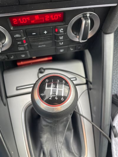 This is a close-up, slightly angled shot of a car's interior, focusing on the center console. The primary subjects are a manual transmission gear shifter and climate control buttons. The gear shifter, a black knob with a chrome and red trim, displays a six-speed manual gear pattern (1, 2, 3, 4, 5, 6, and R for reverse). It appears to be in neutral or a similar position.

To the left of the gear shifter are the climate control elements. A digital display shows "21.0°C" in red, indicating the temperature. Below this are several buttons with icons and text, including controls for temperature adjustment (+/-), fan speed, air conditioning (AC), and auto mode. A silver dial with a black grip is also visible, likely for temperature control.

The background shows the textured fabric of the car's upholstery and a portion of the dashboard. A black cable is visible running across the console, possibly for charging a device or connecting to the infotainment system. The overall scene suggests the interior of a modern passenger car.

There are no people visible in the image. The lighting suggests it might be daytime, but without external views, the weather and precise time of day are indeterminate. The location context provided is Criuleni, Moldova, which, while not directly evident from visual cues within the image, provides a geographical setting. No strong emotions are conveyed; the image is factual and observational. Visible text includes "AUTO", "AC", and the temperature reading "21.0°C". The condition of the gear shifter suggests it has been used, with some minor scuffing visible on the chrome ring.
