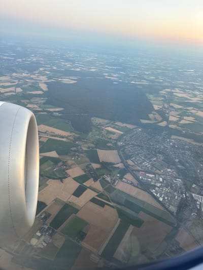 An aerial view shows the city of Bottrop, Germany, during sunset. The image is dominated by the wing of an airplane on the left side, casting a shadow over the landscape. The city is densely populated with buildings, roads, and industrial areas. Surrounding the city are agricultural fields with various crops, separated by hedgerows and small forests. A river runs through the city, and a highway is visible, connecting different parts of the region. In the distance, wind turbines dot the horizon, indicating the presence of renewable energy infrastructure. The sky exhibits a gradient of colors, transitioning from a pale yellow at the horizon to a soft blue overhead, characteristic of a sunset. The overall scene depicts a developed urban area harmoniously integrated with its surrounding rural environment.