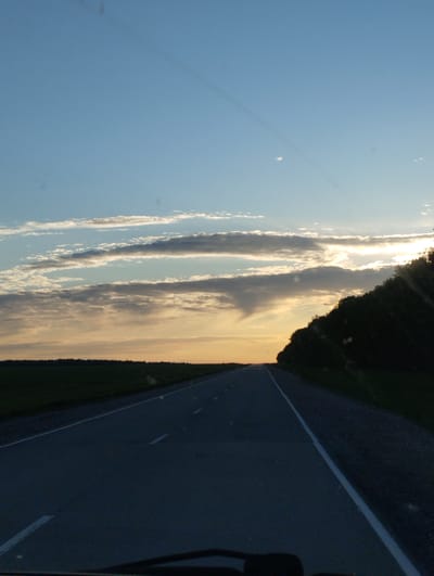 A photograph shows a straight, paved road stretching into the distance. The road is lined with a white stripe on either side, and the pavement appears to be gray. To the left of the road is a flat, green field, and to the right is a line of trees. The sky is a gradient of blue at the top, fading into orange and yellow near the horizon. Wispy clouds are scattered across the sky. The scene is reminiscent of a drive at sunset. The image was taken in Filippovo, Russia.