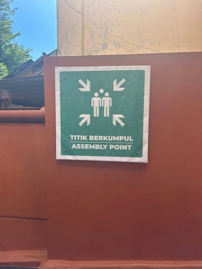 The image shows a green sign with white text and graphics mounted on a reddish-brown wall. The sign indicates an assembly point. The graphics consist of four white arrows pointing inward towards a stylized depiction of three people. The text on the sign reads "TITIK BERKUMPUL ASSEMBLY POINT". The sign is attached to the wall with two visible silver screws.

The wall appears to be textured and painted, with some signs of age or weathering. To the left of the sign, a portion of a pale yellow structure is visible, also showing wear and tear. Below this, a clear blue sky and lush green trees can be seen, suggesting it is daytime and the weather is fair. The overall setting seems to be outdoors, possibly in a public area or compound, given the presence of the assembly point sign. The image is taken from a slightly upward angle, with the sign prominently featured in the center.