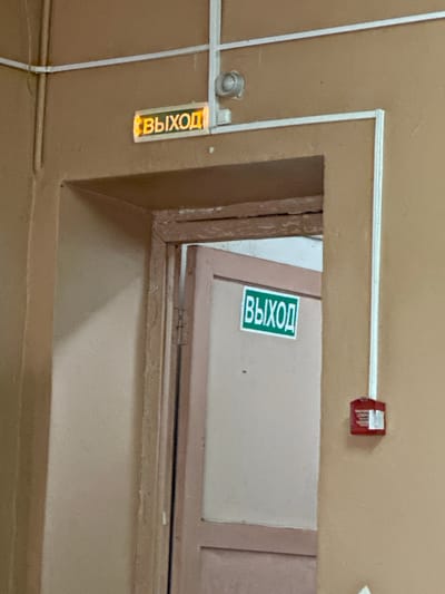 The image shows a hallway with two illuminated exit signs. One sign, located above a doorway, displays the word "ВЫХОД" (Exit) in yellow lettering on an orange background. Below this sign, the doorway is partially open, revealing a door with another "ВЫХОД" sign in green lettering on a white background. To the right of the doorway, a red fire alarm pull station is mounted on the tan-colored wall. The hallway appears to be inside a building, potentially a public space or institution. The lighting is dim, and the walls are painted a neutral color. The overall impression is one of an ordinary interior corridor. The image was taken in Gavrilov Posad, Russia, suggesting the context of a building within that city.