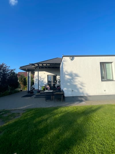 The image depicts a bright, sunny day in a suburban setting, likely in Мухавецкі сельскі Савет, Belarus. The scene features the exterior of a modern, white, two-story house with a dark-tiled roof. A covered patio area extends from the house, furnished with a table and chairs, suggesting an outdoor seating arrangement. The patio is paved with light-colored interlocking stones, which also form a pathway leading around the house.

Adjacent to the house and pathway is a meticulously maintained, vibrant green lawn, casting long shadows from unseen trees or structures, indicating it is likely late afternoon or early morning. The sky is a brilliant, cloudless blue, further emphasizing the clear weather. A single window with blinds is visible on the upper floor of the house, and an outdoor light fixture is mounted on the wall near the roofline.

Vegetation is present, with some trees and bushes visible in the background, adding to the residential feel of the location. The overall impression is one of a tranquil, well-kept residential property on a beautiful day. No people are visible in the image, and there is no visible text.