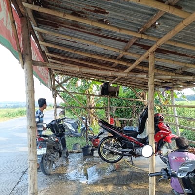 The image depicts a scene in Kecamatan Brangsong, Indonesia (latitude: -6.953, longitude: 110.22). It shows a man, wearing a grey and white striped shirt, standing next to a red motorcycle with a black license plate that reads "6576 QJ". He is looking towards a red and black motorcycle parked beside him. The second motorcycle has a black license plate and a black exhaust pipe. The motorcycles are parked under a thatched roof structure made of bamboo. The roof is supported by wooden poles and has a corrugated metal roof. There is a third, red and black motorcycle in the background, partially obscured by a green plant. Another man, wearing a light blue shirt, is sitting on the red motorcycle, appearing to be working on the engine. The scene is outdoors and there is a paved road leading to the background, where green fields are visible.
