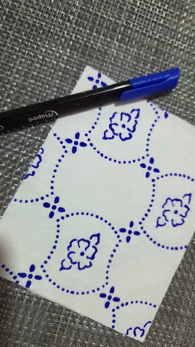 A close-up shot shows a white napkin decorated with a blue floral and dotted pattern resting on a textured gray surface. A blue Maped brand marker is positioned near the napkin. The setting appears to be indoors, likely a home. No people are visible. There is no indication of the location being in Fraccionamiento Real del Valle, Mexico.