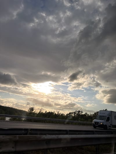 The image captures a roadside view near Kläppa, Sweden, on a cloudy day with the sun breaking through the clouds. A white cargo van with " Rydbergs Trävaror" signage on its side is traveling on a highway. The van is on the right side of the frame, moving away from the viewer. The highway is bordered by a metal guardrail in the foreground and a more substantial guardrail separating the road from a wooded area. The background features a dense forest line along a hill, with trees and vegetation. The sky is filled with dramatic, heavy clouds, but a bright patch of sunlight is visible in the upper left quadrant, illuminating the scene with a warm glow. The overall impression is a snapshot of a journey through a natural landscape under a dynamic sky.