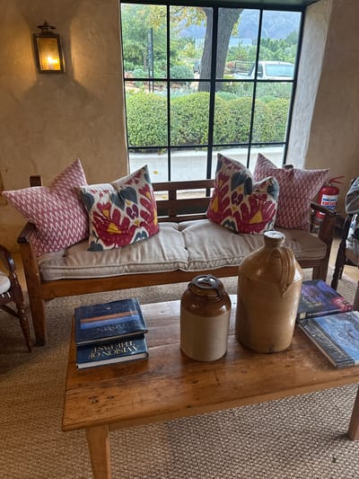 The image shows a well-lit interior in Stellenbosch, South Africa, likely a shop or home, with a focus on a rustic wooden bench situated in front of a large window. The window offers a view of lush greenery and a road. The bench is adorned with patterned pillows, including a geometric pink and white pillow and a floral patterned pillow. On the bench's wooden surface rest two stoneware jugs and two books. One book is titled "A Vision of the Past" and the other book is titled "TRAVINI". In the background, there is a red fire extinguisher. The scene is captured from a slightly high-angle perspective. A decorative lamp can be seen on the left wall. The room has a warm, inviting ambiance due to the natural light and earth-toned decor.