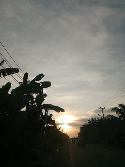 The image depicts a road in Kruengseupeng, Indonesia, at sunset. A car and a motorcycle are traveling on the road. Utility poles and banana trees line the road. The sky is overcast with a visible sun on the horizon.