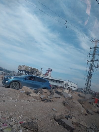 An outdoor scene in a-Zawayda, Palestinian Territory, is depicted under a sky with scattered white cirrus clouds. In the midground, a blue Hyundai Tucson SUV is positioned on uneven terrain composed of mounds of dirt, broken concrete, and asphalt debris. A person's head and shoulder are visible in the driver's seat, oriented towards the right side of the vehicle. In the background, a large, light-colored industrial building displays Arabic script on its side, reading "مصنع غبانة" (Ghabbaneh Factory). Adjacent to this, a multi-story building with a darker facade is partially visible. On the far right, a tall metal lattice transmission tower stands prominently. Overhead power lines with an attached dark, V-shaped object span the upper portion of the frame. The immediate foreground consists of rough terrain composed of shattered asphalt, concrete fragments, and soil.