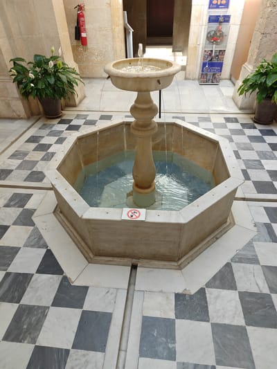 This image depicts an octagonal water fountain with a tiered stone pedestal in a courtyard in Seville, Spain. The fountain is situated on a floor tiled with a checkered pattern of white and dark grey marble squares. To the left of the fountain, a potted plant with large green leaves and white flowers, likely a peace lily, sits on the floor. To the right, another similar potted plant is visible. In the background, a red fire extinguisher is mounted on a stone wall. Further in the background, there is a display stand with colorful graphics and text, including "CATEDRAL DE SEVILLA", suggesting the location is within the Cathedral of Seville's complex. The fountain itself has water flowing from its upper basin into the lower octagonal basin, creating ripples. A small, circular sign with a red border and a line through a drinking glass symbol is affixed to the edge of the fountain's basin. The environment appears to be an indoor or semi-covered courtyard, with stone pillars and arched openings visible. The lighting is somewhat dim, with natural light filtering in from above.