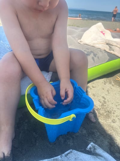 The image shows a close-up view of a child's hands interacting with a blue toy bucket filled with water. The child is wearing dark swim trunks and is sitting on dark sand. A bright green object, possibly a pool noodle, is partially visible behind the child. In the background, the sea and a beach are visible, with a few people in the distance. The lighting suggests it is daytime, likely sunny, given the beach setting. The overall scene depicts a child playing at the beach. There is no visible text in the image.
