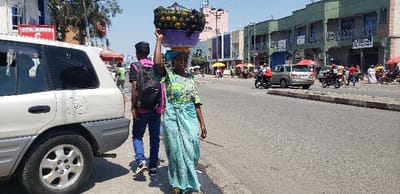 The image shows a woman walking down a street in Rubavu, Rwanda. She is carrying a large basket of fruit on her head. The fruit is likely pineapples, which are a common staple in the region. The woman is wearing a blue dress and a headscarf. Behind her is a man wearing a pink shirt and a black backpack. He is walking alongside a white SUV parked on the side of the road. There are other people walking in the street, and several cars and motorbikes parked along the roadside. The street is lined with buildings on both sides, with several storefronts and a sign that reads "ETS AMOS." The sky is clear and blue. This location is situated near the border of the Democratic Republic of Congo.