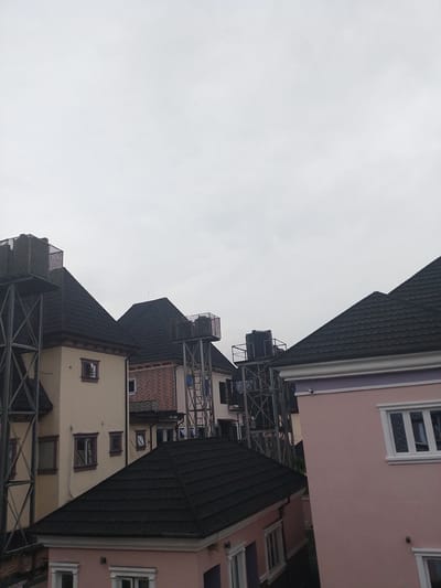 The image depicts a cluster of modern buildings in Rumuomasi, Nigeria, under an overcast sky. The scene is characterized by multiple structures with dark, sloped roofs, suggesting a residential or commercial development. Several of the buildings are equipped with large water tanks supported by sturdy metal scaffolding, a common feature in areas where reliable municipal water supply might be inconsistent. One prominent building is painted a soft pink with white-trimmed windows. Another adjacent structure features a facade of red brick. The overall impression is one of ongoing construction or recent development, with the metal structures indicating infrastructure for water storage. The weather appears to be cloudy and possibly humid, typical of a tropical climate. No people are visible in the frame, and the focus is on the architectural elements and infrastructure of the setting. The time of day is indeterminate due to the uniform lighting from the overcast sky.