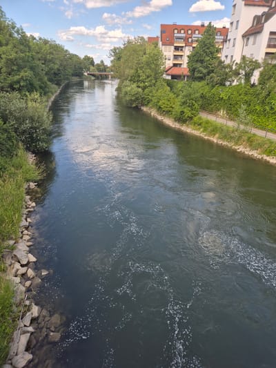 The image shows a wide river flowing through a city, with buildings and greenery on either side. On the right bank, there are modern apartment buildings with red-tiled roofs and white facades, set against a backdrop of lush green trees and ivy-covered walls. A paved walkway runs along the riverbank, bordered by a metal railing. On the left bank, dense green trees and shrubs line the water's edge, with large rocks and grass creating a more natural embankment. The river itself is dark blue-green, with ripples and small white foam patches suggesting a gentle current. In the distance, a bridge with an orange railing crosses the river, and more trees and buildings can be seen along the banks. The sky is blue with scattered white clouds. The scene appears to be a view from a bridge or an elevated vantage point overlooking the Isar River in Landshut, Germany. There are no people visible in the image.