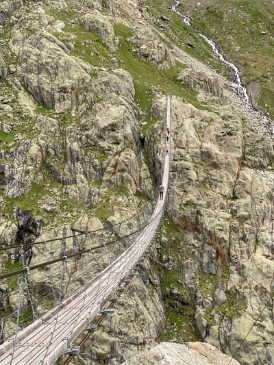 This image captures a long pedestrian suspension bridge stretching across a rugged, mountainous valley in Innertkirchen, Switzerland. The bridge, constructed with wooden planks for the walkway and robust metal cables and vertical mesh for support and railings, spans between two steep, rocky slopes.

Several individuals are visible traversing the bridge. In the mid-ground, a person wearing a grey top and dark trousers walks towards the left side of the frame. Further along the bridge, a small group of three individuals can be seen, distinguishable by their clothing: one in yellow, one in black, and one in a light-colored top. At the far end of the bridge, near its anchor point on the distant cliff face, a single figure in orange is partially visible. All individuals appear to be walking in the same direction, from right to left across the bridge.

The surrounding environment consists of densely packed, large grey rock formations interspersed with patches of vibrant green vegetation, primarily grass and moss. The terrain is steep and appears challenging. In the upper right quadrant of the image, a narrow waterfall or stream cascades down a rock-strewn slope, flowing from the higher ground towards the unseen depths below. The lighting is consistent, suggesting an outdoor daylight setting without direct harsh sunlight.