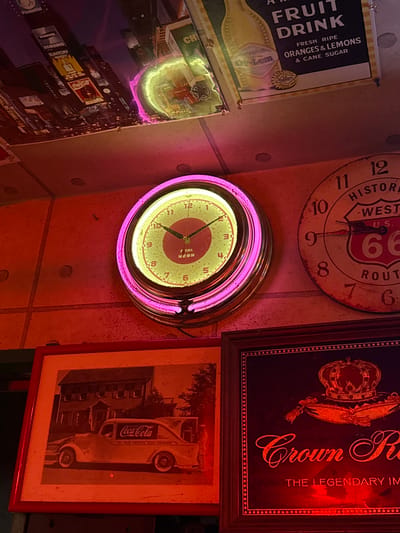 The image shows a collection of vintage-themed items displayed on a wall, likely in a bar or diner setting, possibly in Adachi, Japan. The main subjects are three framed pieces and a clock. At the top left, a framed sign advertises "FRUIT DRINK" with "FRESH RIPE ORANGES & LEMONS & CANE SUGAR." Below this, a framed poster depicts a nighttime cityscape with numerous illuminated billboards, including advertisements for Coca-Cola, Samsung, and Suntory Whisky, suggesting a Times Square-like environment. To the right of the cityscape poster is a framed advertisement for "Crown Royal," featuring a crown and text indicating "THE LEGENDARY IM."
Dominating the center of the image is a round clock with a neon pink outer rim. The clock face has a pinkish-yellow glow and displays numbers from 1 to 12, with hands indicating a time around 9:50. Below this clock, another circular object, possibly a sign or a smaller clock, has a neon green glow. A larger, distressed round clock face is visible at the top center, featuring route 66 imagery and historical markings. A framed advertisement for Coca-Cola with a classic truck is situated at the bottom right. The lighting is warm and atmospheric, with red and pink neon tones prevalent, creating a retro ambiance. The overall impression is one of nostalgia and Americana, perhaps curated in a Japanese establishment. No people are visible in the image.