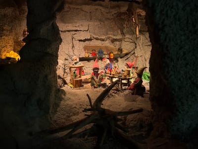The image depicts a scene from the Fairytale Forest (Märchenwald) in Tolk, Germany. It is an indoor exhibit designed to resemble a cave dwelling. In the center of the scene, a group of five animatronic gnomes are gathered around a rustic wooden table. Four of the gnomes are actively engaged in activities. One gnome, wearing a red conical hat, is seated and appears to be reading a book. Another gnome, also with a red hat, is standing and holding a ladle. A third gnome, in a green pointy hat, is seated on a stool. The fourth gnome, wearing a blue shirt and red pants, is standing and reaching towards the table. The fifth gnome, partially obscured by a structure on the left, is visible from the shoulders up.

The environment is designed to look like a cave with rough, textured walls. The floor is covered in sand and small stones. A prominent piece of gnarled driftwood lies in the foreground. Behind the gnomes, a wooden shelf is mounted on the cave wall, displaying various miniature household items such as a red teapot, a blue pot, and a lantern. The lighting is dim and atmospheric, with warm yellow and orange tones illuminating the scene, suggesting an interior, underground setting. The overall impression is one of a cozy, albeit slightly eerie, gnome community.