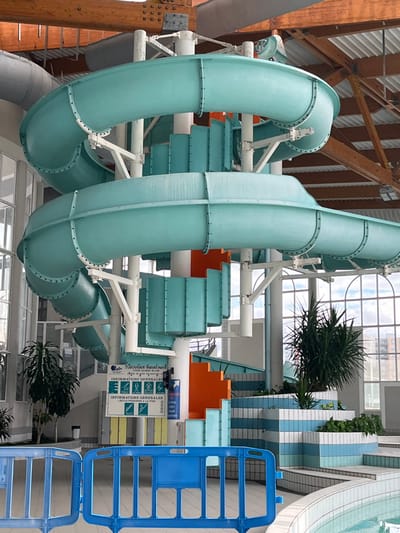 The scene is an indoor aquatic center located in Andrézieux-Bouthéon, France. A prominent teal-colored enclosed spiral water slide, constructed from segmented plastic sections, dominates the center of the frame, supported by white tubular columns and metal framework. An integrated spiral staircase, featuring orange treads and teal-colored risers, ascends alongside the slide. At the base of the structure, a white sign with French text titled "TUBOGGAN AQUATIQUE" and "INFORMATIONS GENERALES" displays safety pictograms, including a rule for '1 SEULE PERSONNE À LA FOIS' (1 person at a time). The facility features a high, pitched ceiling with exposed wooden trusses and white corrugated panels. Large arched windows along the right wall provide natural light and a view of distant urban buildings under a cloudy sky. The pool area in the foreground exhibits stepped edges with light blue and white striped ceramic tiles. Potted green plants are strategically placed around the base of the slide and within the tiled pool environment. A blue plastic safety barrier is positioned horizontally across the lower foreground, partially obscuring the pool. No individuals are present within the facility.