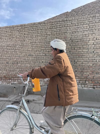 A male individual is depicted riding a silver-colored bicycle in Nakhonay, Afghanistan. He wears a white turban over a white mesh cap, a brown padded jacket with black zippered pockets, light-colored baggy trousers, and white athletic shoes. In his right hand, he holds a yellow plastic jug or container with a visible red label. The bicycle features front and rear fenders and a metal rack over the rear wheel. The background consists of a high, weathered brick wall constructed from light-colored bricks. Above the wall, a clear blue sky with sparse clouds is visible. The ground beneath the bicycle appears to be an uneven concrete or packed earth surface. The scene is brightly lit by natural daylight.