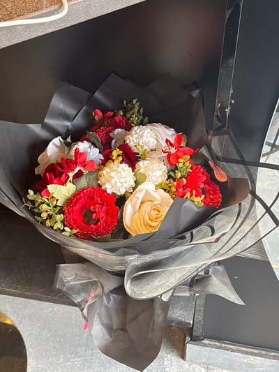 A bouquet of artificial flowers is displayed on a dark, textured surface. The bouquet features a variety of blooms in red, white, and cream colors, arranged with green faux foliage and wrapped in black paper and translucent cellophane. The backdrop is dark and appears to be a wall or shelf. The lighting is moderate, casting soft shadows. The image is a close-up shot of the bouquet. The location is identified as Sweileh, Jordan.